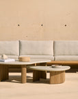 Minimalist outdoor seating area with a light beige cushioned sofa and two Ethnicraft Boomerang Outdoor Coffee Tables, each featuring hand-finished concrete tops and ribbed reclaimed teak bases, set against a smooth beige wall with small holes.