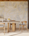 A modern dining area showcases the Ethnicraft Trestle Outdoor Dining Table with chairs, a bowl of lemons, ceramic cups, and a pitcher, set against a beige stone wall beneath a wooden ceiling.