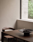 A minimal dining area with a wooden table and chair displays Ethnicraft’s Satellite Bowl filled with dark cherries. Sunlight streams through the window, casting shadows on neutral walls reminiscent of Ethnicrafts design style.