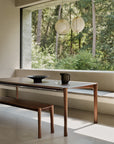 Modern dining area with Ethnicraft's Pi Dining Table - Teak Brown, built-in bench seating, a black pitcher and bowl, a large forest-view window, and two round pendant lights above.