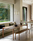 A modern dining area featuring Ethnicraft’s Pi Bench in teak with cushions, paired with a wooden table, accented by a bowl and pitcher, large windows framing greenery, and pendant lights for classic European style.