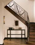 A wooden staircase with black metal railings curves above a minimalist Ethnicraft PI Console in dark brown teak. Decorative accents and abstract framed art enhance the space’s neutral palette.