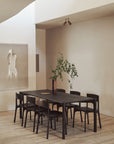 Minimalist dining room with an Ethnicraft PI Dining Table in dark brown teak, eight matching black chairs, a tall vase with green branches, light-colored walls, wood flooring, and abstract wall art.