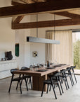 A modern dining room features Ethnicraft’s Double Extendable Dining Table in natural teak set for eight, black chairs, minimal tableware, a linear pendant light above, exposed wooden beams, and neutral curtains letting in natural light.