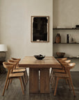 A minimalist dining room features an Ethnicraft wooden table and eight Bok Dining Chairs by Ethnicraft in natural teak, a dark bowl centerpiece, neutral walls, shelves with pottery and books, and abstract black artwork above the table.