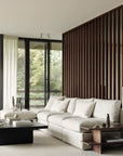 A modern living room features a large beige sectional sofa, an Ethnicraft Kabuki Side Table in varnished teak, a wooden slat room divider, sheer curtains, and large windows overlooking greenery.