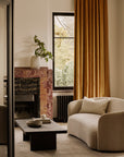 A cozy living room features a curved beige sofa, Ethnicraft’s Grooves Coffee Table, and a marble fireplace with a white vase and greenery. Gold curtains frame a large window, filling the space with natural light for a warm, inviting feel.