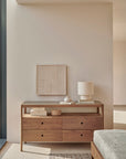 The Ethnicraft Spindle Dresser in reclaimed FSC teak displays books, decorative boxes, a vase, and a textured lamp. A neutral abstract artwork hangs on a light beige wall in a bright, modern room.