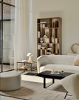 A modern, minimalist living room features neutral-toned furniture, a curved sofa, a round ottoman, and a black coffee table atop the Ethnicraft Dunes Rug - Cumin. Sunlight pours in through large windows onto the tall wooden bookshelf.