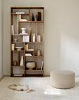 A minimalist room features a tall wooden bookshelf with books and neutral vases, beside an Ethnicraft Barrow Pouf - Off White / Large upholstered in Italian fabric on a light rug. Wooden toy train tracks and trains lie on the floor.
