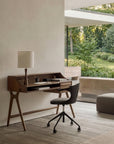 A modern workspace featuring the Ethnicraft Flow Desk, paired with a lamp, chair, and stationery, sits by a large window overlooking a lush green garden. Natural light fills the minimalist, serene room with neutral tones and simple decor.
