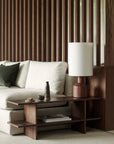 A modern living room includes a beige sofa with a dark green pillow and an Ethnicraft Kabuki Side Table in varnished teak, topped with a lamp, small vase, and snack, set before vertical wooden slats.