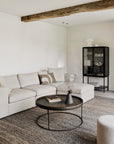 A modern living room featuring a beige sectional sofa, Ethnicraft Tray Coffee Table, textured rug, and a glass cabinet with decor against a light wall. An exposed wooden ceiling beam completes the space.