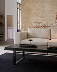 A modern living room with European refinement features a beige sofa, the Ethnicraft Stability Coffee Table - Umber topped with a wine glass and tray, a tall floor lamp, an exposed brick wall, and a large window inviting natural light.