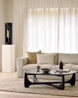 A modern living room features the Ethnicraft Mellow Sofa in off white, beige and white cushions, a textured rug, sculptural black coffee table, sheer white curtains on a large window, and a black abstract sculpture on an Ethnicraft pedestal.
