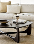 A modern living room with a beige sofa, cream and tan pillows, and an open book. The Ethnicraft PI Coffee Table—Mahogany, Pebble Shape—displays a bowl, books, decor, and plant atop a light textured rug.