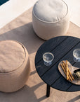 Two round beige ottomans sit on an Ethnicraft Nomad Outdoor Rug beside a round black wooden table holding two glasses of white wine and a platter with breadsticks and dip.