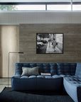 A modern living room features the Ethnicraft N701 Sectional Sofa in blue, a black floor lamp, textured wall, and a black-and-white photo above the sofa. Light pours in through a high horizontal window.