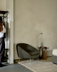A dark gray Ethnicraft Barrow Lounge Chair - Paon with metal legs sits on a rug next to a small wooden side table with a carafe and a glass.