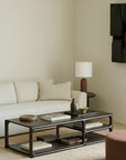 A minimalist living room featuring a light beige sofa, a neutral pillow, the Ethnicraft Eda Coffee Table adorned with books and decor, a round mahogany occasional table with a brown vase lamp, and abstract wall art—all showcasing heirloom-quality furniture throughout the space.