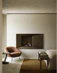 A minimalist bedroom features an Ethnicraft Bok Bench in varnished teak, a modern fireplace, rust-colored lounge chair, side table, books and wooden accents, beige bed, and brown rug on a light floor for a clean, neutral look.