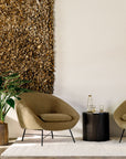 A modern, cozy seating area with two tan armchairs, a round black side table, a leafy plant, textured brown wall art, and the Ethnicraft Dunes Rug - Sand hand-knotted using the Persian technique completes the neutral background.