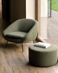 A modern olive armchair stands beside the Ethnicraft Barrow Pouf - Pine / Large, topped with books near a window facing a patio and lawn. The wood-floored room features quality Italian fabrics.