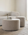 A minimalist living room with a curved beige sofa, round textured ottoman, and matching side table sits atop the Ethnicraft Dunes Rug - Cumin. Two glass bottles and two glasses are arranged on the ottoman.