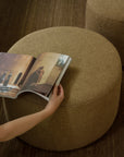 A hand holds open a magazine resting on the Barrow Pouf - Ginger / Large by Ethnicraft, crafted from premium Italian fabrics and set on a brown carpet, with another matching pouf in the background.