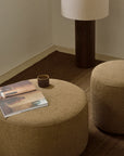 A cozy corner with two Ethnicraft Barrow Pouf - Ginger / Large, a brown floor mat, a wooden lamp with a light shade, a small ceramic cup, and an open magazine on one pouf.