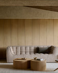 Minimalist living room with neutral tones, featuring the Ethnicraft N701 3-Seater Sofa in beige tufted upholstery, round wooden coffee tables, a sculptural wooden chair, floor lamp, and textured wood panel walls. A light rug covers the floor, highlighting timeless European design influences.