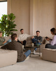 Five people relax in a modern living room on beige sofas and ottomans, chatting over coffee mugs placed on the Ethnicraft Tray Coffee Table - Set of 2, while a large green plant adds to the inviting atmosphere.