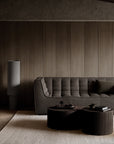 Minimalist living room featuring the Ethnicraft N701 3-Seater Sofa in Dark Grey with a matching pillow, two round coffee tables styled with books and cups, a textured beige rug, and a tall cylindrical lamp set against a paneled wooden wall.