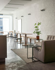 A modern, minimal cafe interior with white brick walls, Ethnicraft Torsion Square Dining Tables in oak, matching chairs, a potted plant, and a cozy gray armchair by a small table with water on a rug. The space is bright with natural light.