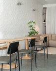 Minimalist cafe interior featuring Ethnicrafts Torsion Square Dining Table in oak, black chairs, a concrete floor, white brick wall, potted plant, and a beige sofa with a black floor lamp by sheer white curtains.
