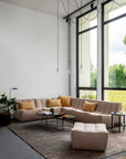 Modern living room with large windows, beige sectional sofa with mustard yellow cushions, black metal base ottoman, Nesting Coffee Table Charcoal - Set of 2 by Ethnicraft, potted plant, floor lamp, exposed bulb lights, and a charcoal mirror above the textured rug.