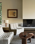 A modern living room features the Ethnicraft N701 Modular Sofa in Ecru Eco Fabric, two tufted beige chairs, a wooden coffee table with glasses and bowls, a rug, fireplace, table lamp, abstract art, and a large window overlooking green foliage.