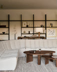 A modern living room features Ethnicraft's N701 Modular Sofa in ecru eco fabric, two nested wooden coffee tables, bookshelves with decor, framed art, a lamp, and neutral accents—creating a minimalist space that’s inviting and easy to maintain.