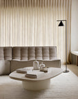 A modern living room features the Ethnicraft N701 Modular Sofa in ecru eco fabric, a white oval coffee table with books and vases, a black floor lamp, and sheer beige curtains that filter soft natural light.