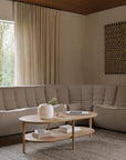 A neutral living room with a beige sectional sofa and an Ethnicraft Bok Coffee Table in oak, styled with a white vase, books, and a tray, complemented by textured curtains and wall art in the background.