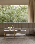 A modern living room featuring the Ethnicraft N701 Modular Sofa in Ecru (Eco Fabric), a round wooden coffee table with books and a potted plant, neutral carpet, large window with beige curtains, and green trees outside.