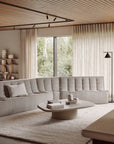 A modern living room featuring the Ethnicraft N701 Modular Sofa in ecru eco fabric, a round coffee table with books and decor, a soft beige rug, floor-to-ceiling curtains, and a window overlooking greenery.