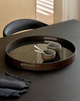 The Ethnicraft Second Nature Tray, featuring a dark wood finish with a circular pattern, holds two ceramic cups and a spoon on a black table beside a chair, with folded cloth nearby and sunlight filtering through blinds.