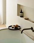 A minimalist bathroom features a white tub filling with water, a bronze faucet, and an Ethnicraft Second Nature Valet Tray holding a sponge and bar soap. A shelf above displays a pump bottle and brush against a neutral-toned wall.