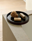 A black Second Nature Valet Tray by Ethnicraft holds a natural sea sponge, wooden nail brush, and bar of soap on the stone tub’s edge, with light wood flooring in the background.