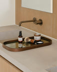 A bathroom counter with a modern wall-mounted faucet and an Ethnicraft Second Nature Valet Tray holding shaving cream, a brush, a small bottle, and an open tin—all reflected in the tray’s silk-screen printed glass surface.