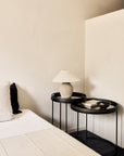 A minimalist bedroom features a white bed with cream and dark pillows, two black Ethnicraft Round Tray Side Tables (S/L), a beige table lamp, stacked books, and a small bowl against light neutral walls.