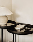 A minimalist interior features the Ethnicraft Round Tray Side Table Set - S/L: one table holds a beige ceramic lamp with a fabric shade, the other displays a closed book and a small black bowl filled with decorative items.