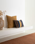 Two decorative pillows—one solid mustard yellow, one brown with blue and yellow stripes—rest on a white bench. Nearby, a woven vase holds dried flowers and an Ethnicraft Nomad Kilim Rug in Terracotta covers the floor.
