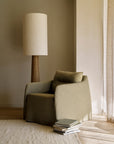 The Ethnicraft Weave Lounge Chair in green linen-blend fabric sits next to a tall, textured floor lamp with a wooden base. Several books rest on a light rug beside the chair in this softly lit room.